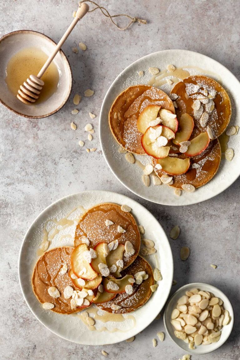 two plates with 3 pancakes each, baked apple slices, almond flakes, maple syrup and icing sugar on top. A small bowl with maple syrup and another small bowl with more almond flakes