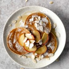 a plate with 3 pancakes, baked apple sliced, almond flakes, maple syrup and icing sugar on top