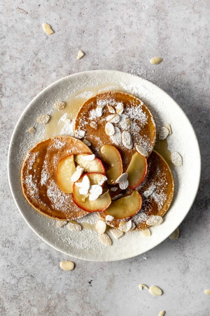 a plate with 3 pancakes, baked apple sliced, almond flakes, maple syrup and icing sugar on top