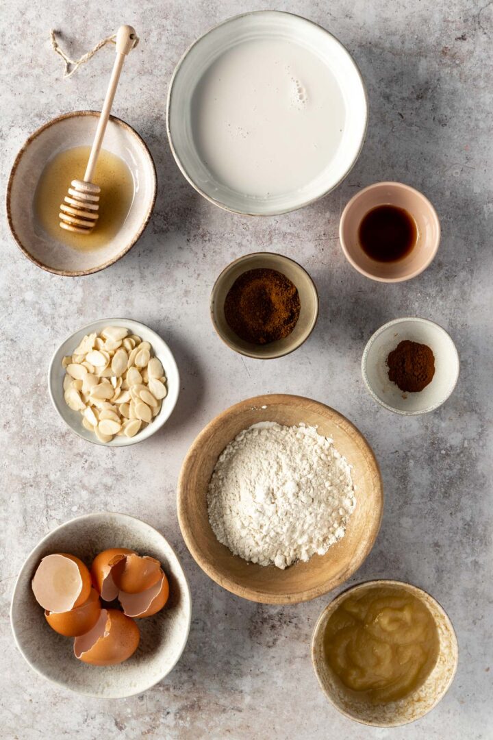 ingredients to make apple pancakes. A variety of bowls with almond milk, maple syrup, coconut sugar, vanilla extract, cinnamon, almond flakes, flour, eggs shells and apple puree
