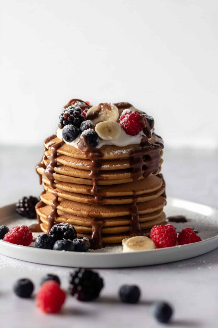 a plate with a stack of pancakes, coconut yogurt, sliced banana, berries, chocolate ganache and powdered sugar on top