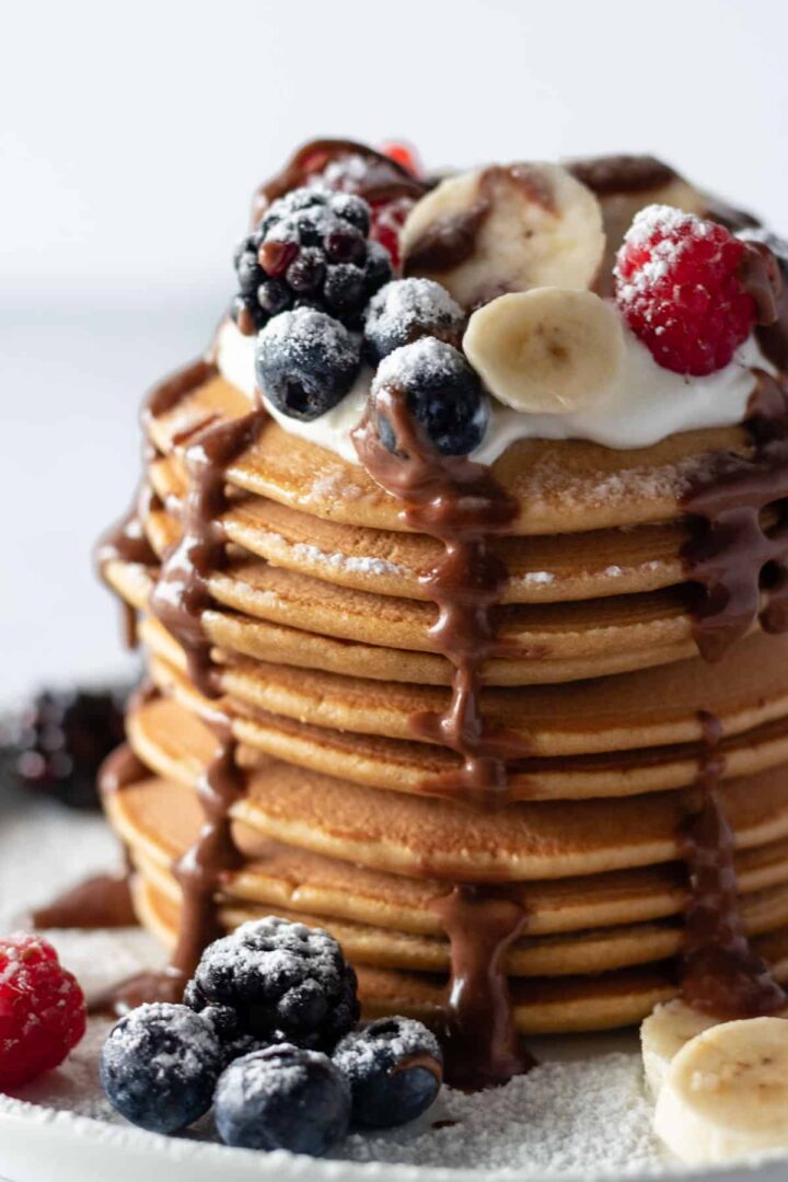 close up of a plate with a stack of pancakes, coconut yogurt, sliced banana, berries, chocolate ganache and powdered sugar on top