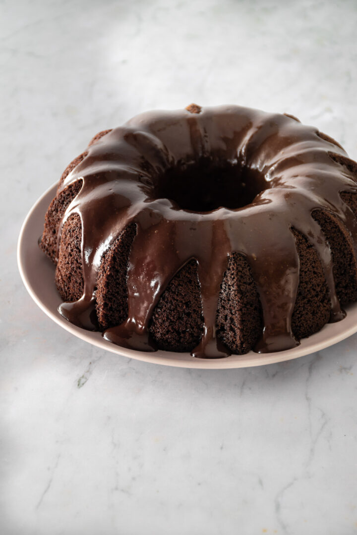 a bundt chocolate cake with chocolate glaze on top on a pink plate on top of a marble table