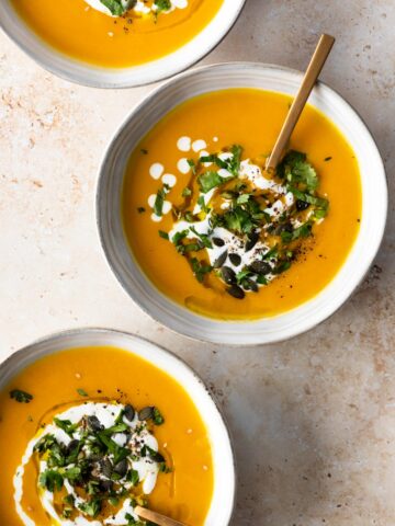 three bowls with soup, a drizzle of coconut milk, pumpkin seeds, a drizzle of olive oil and chopped coriander on top
