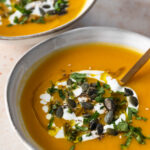 close up of two bowls with soup, a drizzle of coconut milk, pumpkin seeds, a drizzle of olive oil and chopped coriander on top