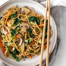 a plate on top of a kitchen towel with noodles, sliced mushrooms, julienned carrots, chopped cabbage, chopped green onions, black sesame seeds on top and chopsticks on the side