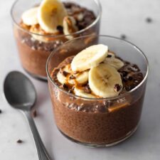 two glass bowls with chocolate chia pudding, sliced banana, cacao nibs, peanut butter and a spoon on the side