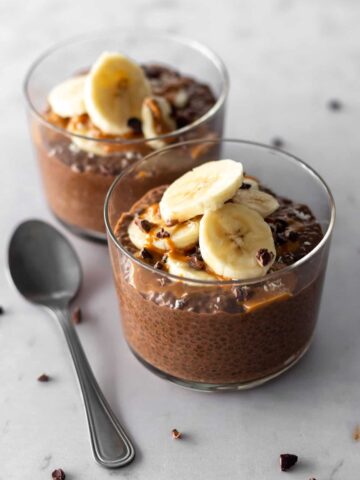 two glass bowls with chocolate chia pudding, sliced banana, cacao nibs, peanut butter and a spoon on the side