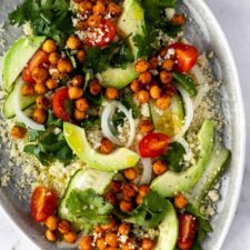 close up of a plate with couscous, sliced avocado, sliced cucumber, cherry tomatoes, roasted chickpeas, sliced onion and coriander