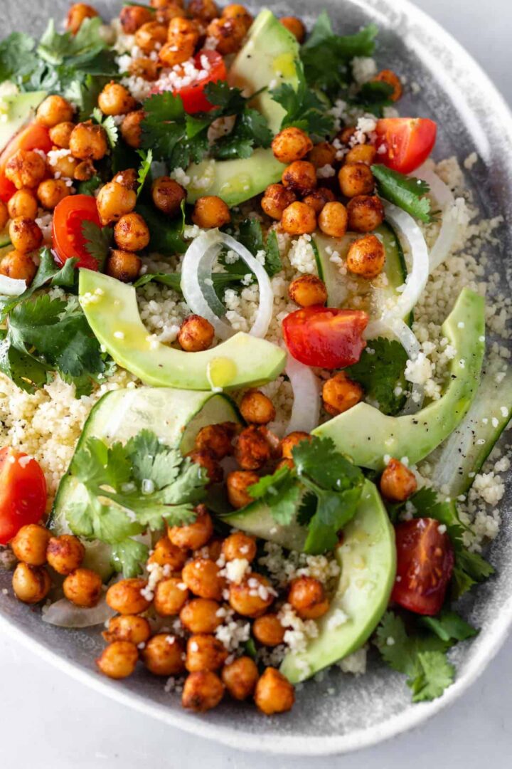 close up of a plate with couscous, sliced avocado, sliced cucumber, cherry tomatoes, roasted chickpeas, sliced onion and coriander