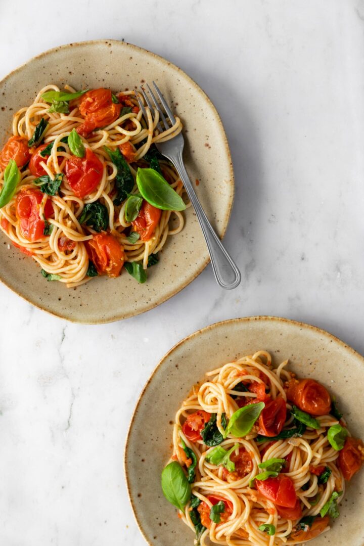 two plates with creamy spaghetti pasta, cherry tomatoes, spinach and basil leaves on top of a marble table