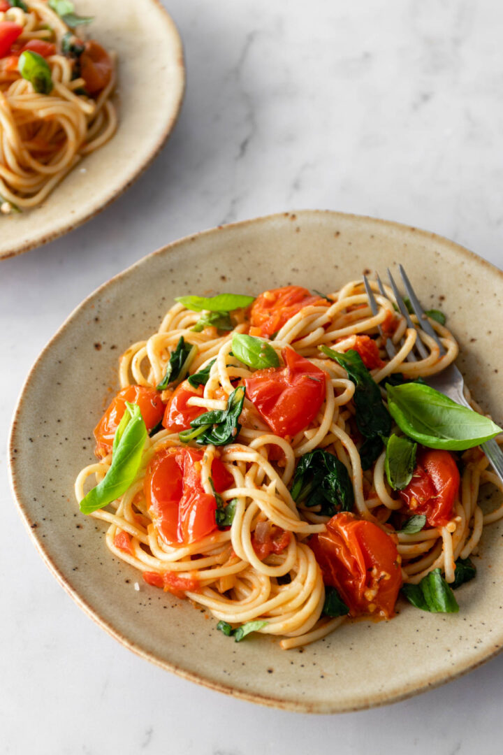 a plate with spaghetti, cherry tomatoes, spinach, basil leaves and a fork on the side. Another plate out of focus on the top left corner