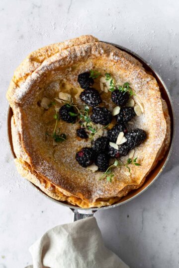 pancake on a skillet with blackberries, thyme, powdered sugar and almond flakes on top