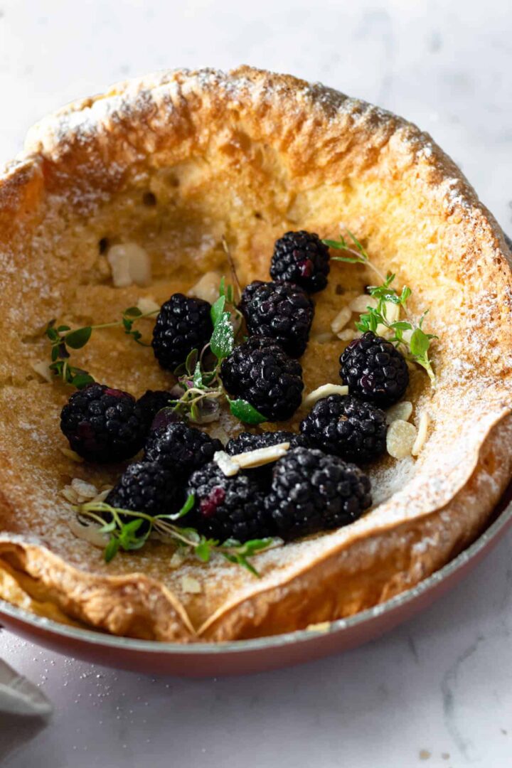dutch baby pancake close up on a skillet with blackberries, almond flakes, thyme and powdered sugar on top