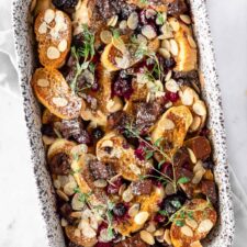 a casserole with sliced bread, chocolate chunks, flaked almonds, berries, icing sugar and thyme on top of a kitchen towel