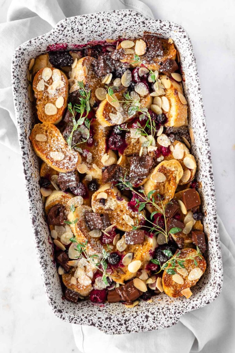 a casserole with sliced bread, chocolate chunks, flaked almonds, berries, icing sugar and thyme on top of a kitchen towel
