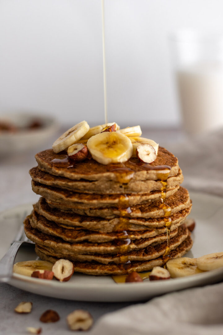 a plate with a stack of pancakes with banana slices, hazelnuts and maple syrup on top