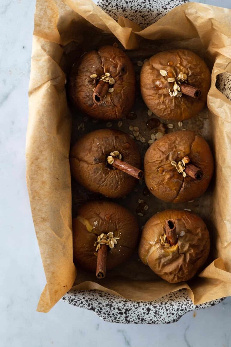 a baking casserole with parchment paper, baked apples with a cinnamon stick oats and maple syrup on top of a marble table