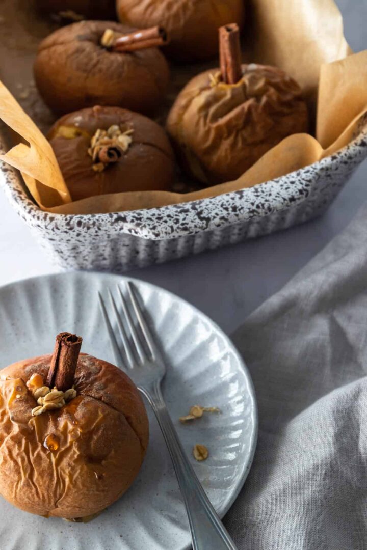 a baking casserole with parchment paper, baked apples with a cinnamon stick oats and maple syrup, a kitchen towel and a small plate with a baked apple and a for on the side