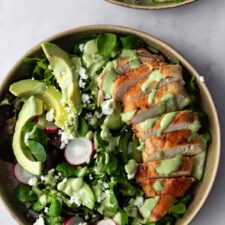 a bowl with sliced chicken breast, sliced avocado, radishes, feta cheese, cucumber, salad mixture and green dressing on top