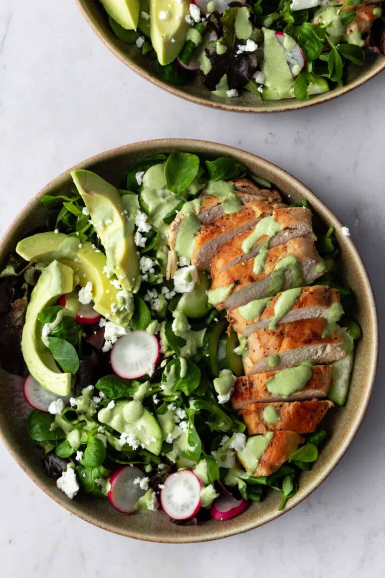 a bowl with sliced chicken breast, sliced avocado, radishes, feta cheese, cucumber, salad mixture and green dressing on top