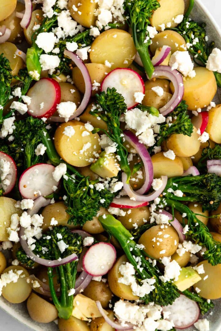 close up of a large plate with a healthy potato salad with new potatoes, broccolini, cubed avocado, red onion, sliced radishes and crumbled feta cheese
