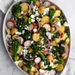 a large plate with new potatoes, broccolini, cubed avocado, red onion, sliced radishes and crumbled feta cheese on top of a grey kitchen towel and a marble table