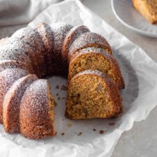 a bundt carrot cake with powdered sugar on top, sliced, on top of white parchment paper on a beige board, two small plates with one cake slice each out of focus