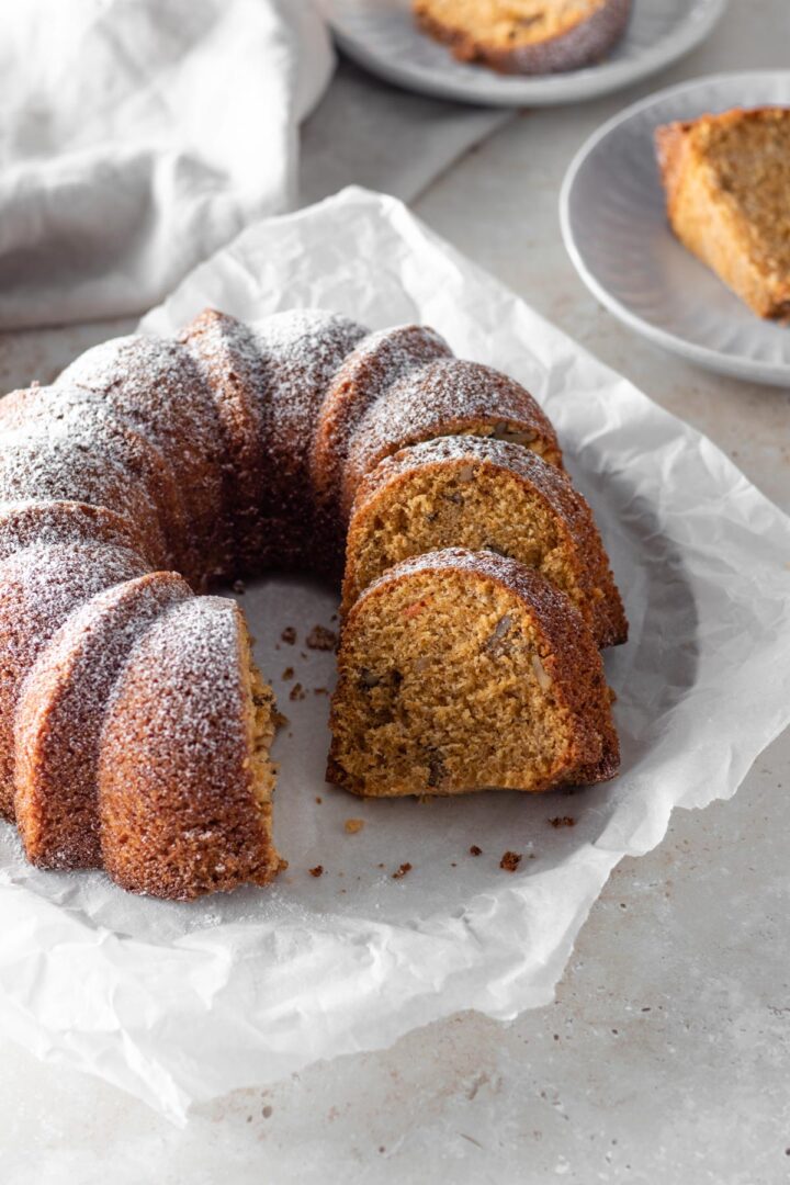 a bundt carrot cake with powdered sugar on top, sliced, on top of white parchment paper on a beige board, two small plates with one cake slice each out of focus