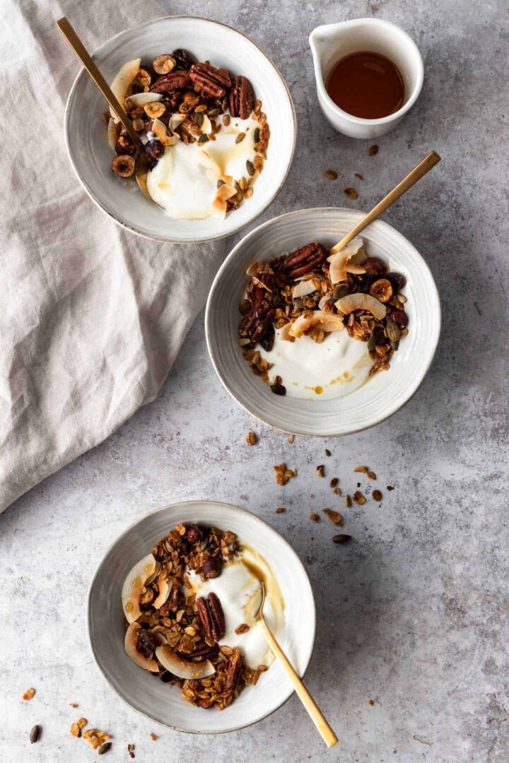 three bowls with granola, coconut yogurt and a small jug with maple syrup