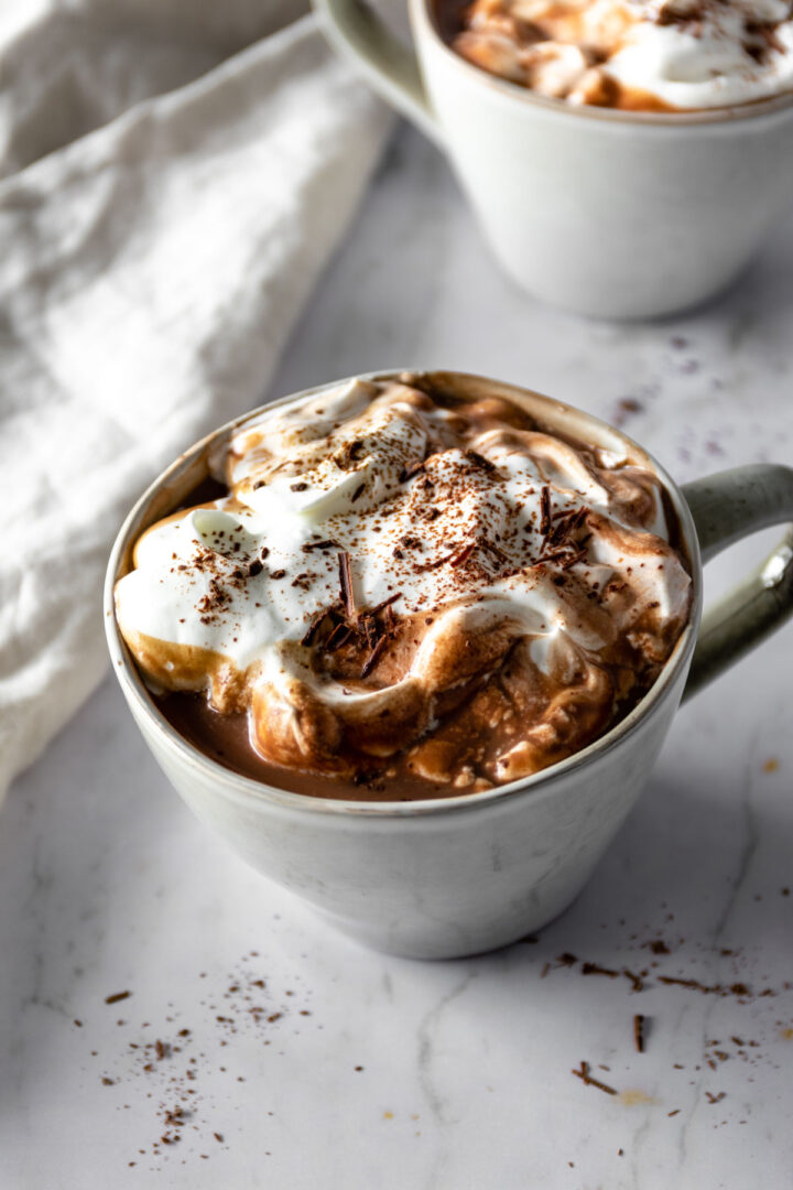 a mug with hot chocolate topped with whipped cream and shredded chocolate on top, another mug out of focus on the top right corner