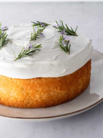close up lemon cake on a plate with whipped cream frosting on top and decorated with rosemary