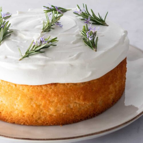 close up lemon cake on a plate with whipped cream frosting on top and decorated with rosemary