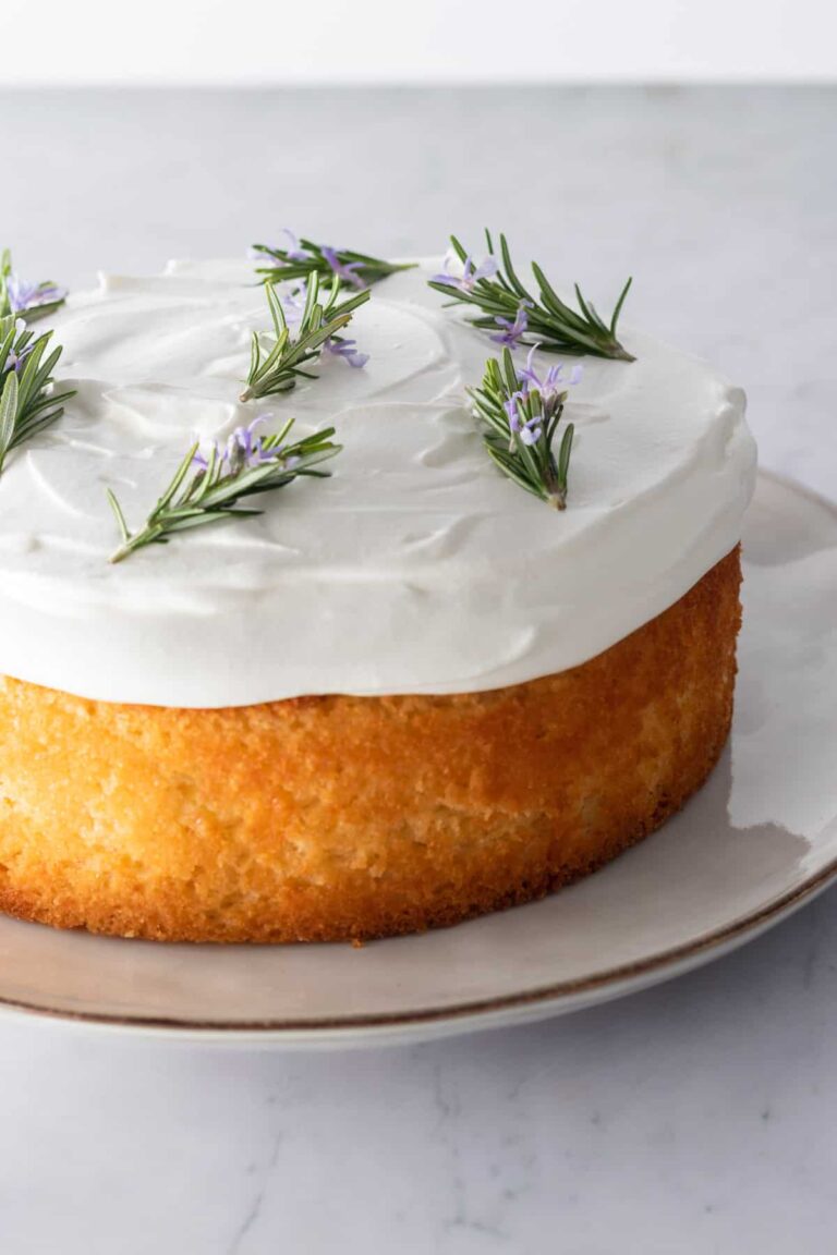 close up lemon cake on a plate with whipped cream frosting on top and decorated with rosemary