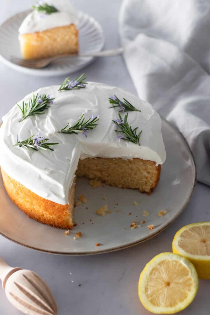 lemon cake on a plate with whipped cream frosting on top and decorated with rosemary. A small plate with a slice of lemon cake on the top left corner, a lemon cut in the middle on the bottom right corner, a grey kitchen towel and a wooden lemon squeezer on the bottom left corner