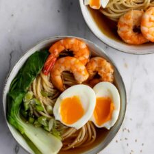 two ramen bowls with noodles, shrimp, boiled egg, green onions, bok choy and a pinch bowl with sesame seeds on the side