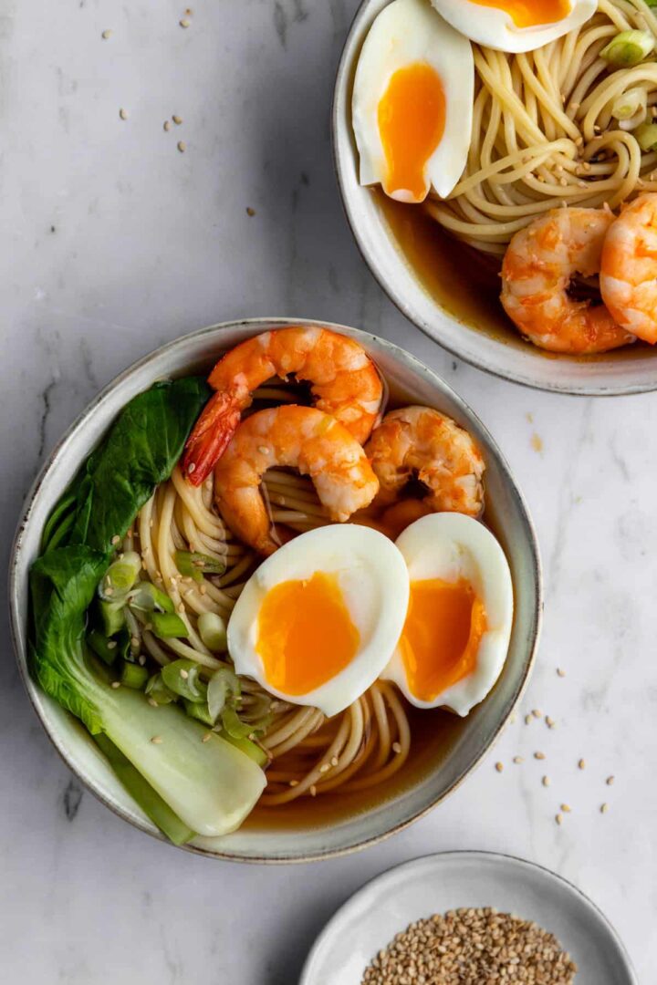 two ramen bowls with noodles, shrimp, boiled egg, green onions, bok choy and a pinch bowl with sesame seeds on the side