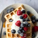 a plate with waffles, blueberries, raspberries, yogurt, maple syrup and icing sugar on top