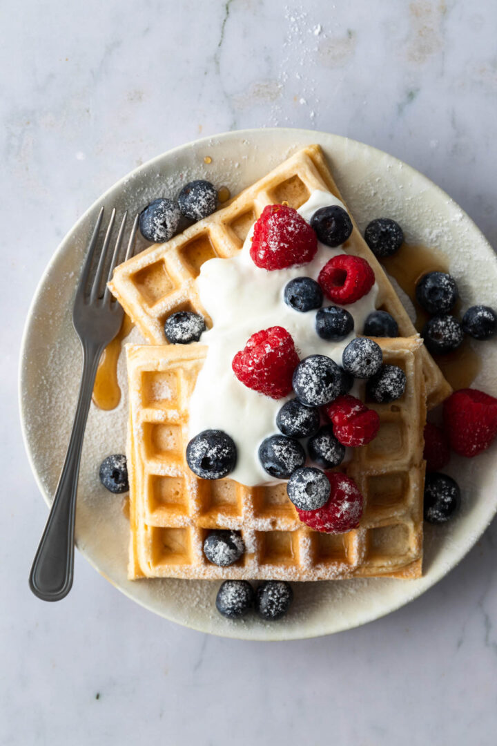 a plate with waffles, blueberries, raspberries, yogurt, maple syrup and icing sugar on top and a fork on the side