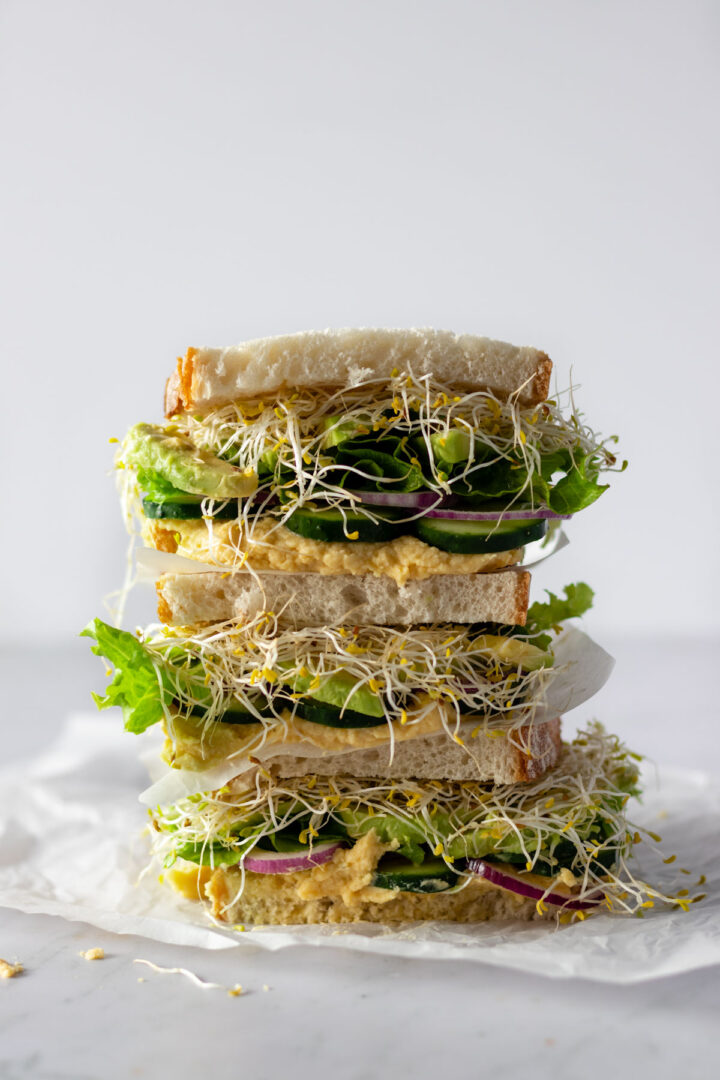 hummus sandwiches with avocado, red onion, cucumber, lettuce and micro greens on top of parchment paper