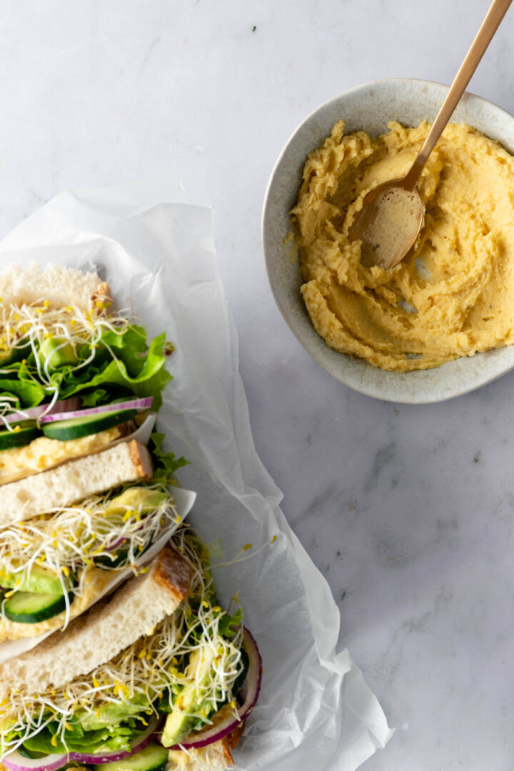 hummus sandwiches with avocado, red onion, cucumber, lettuce and micro greens on top of parchment paper, a bowl with a spoon and more hummus