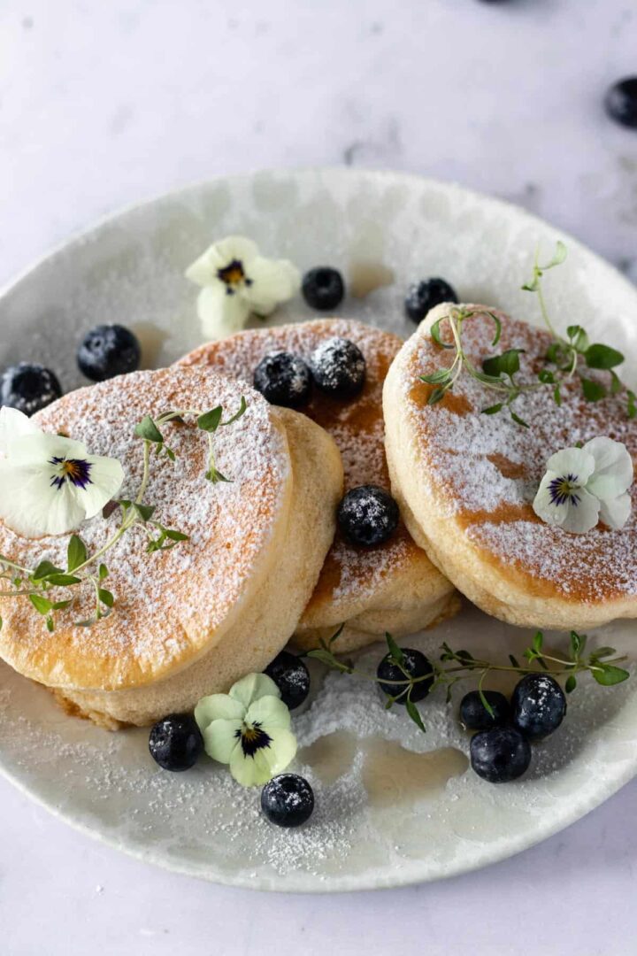 close up of a plate with three pancakes, blueberries, thyme, edible flowers, maple syrup and powdered sugar on top