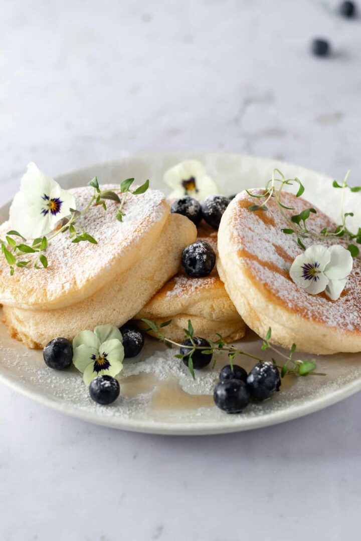 a plate with three pancakes, blueberries, thyme, edible flowers, maple syrup and powdered sugar on top