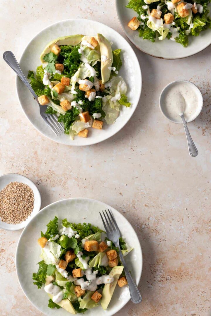 three plates served with iceberg lettuce, kale, bread croutons, avocado, white dressing and sesame seeds on top. A small bowl with a spoon and more dressing on the side and a small plate with sesame seeds