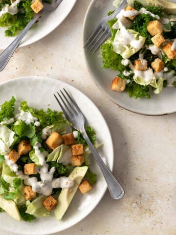 three plates served with iceberg lettuce, sliced avocado, kale, bread croutons, white dressing and sesame seeds on top
