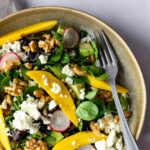 a bowl with mixed greens, cubed avocado, sliced radishes, sliced cucumber, chopped walnuts, mango and crumbled feta cheese on top of a beige kitchen towel
