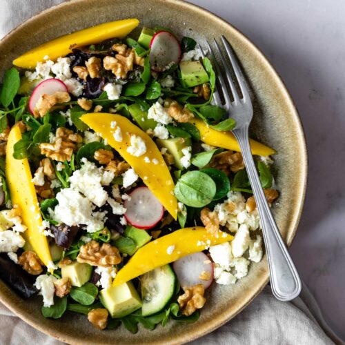a bowl with mixed greens, cubed avocado, sliced radishes, sliced cucumber, chopped walnuts, mango and crumbled feta cheese on top of a beige kitchen towel