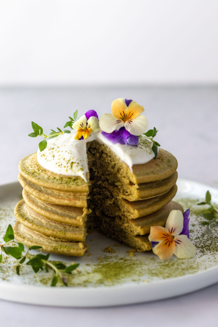 a plate with a stack of sliced pancakes, coconut yogurt, edible flowers and match powder on top