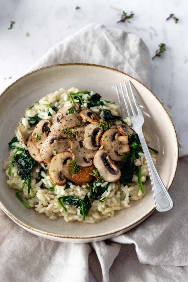 a plate with mushroom risotto with spinach, garnished with thyme and a fork on the side on top of a kitchen towel