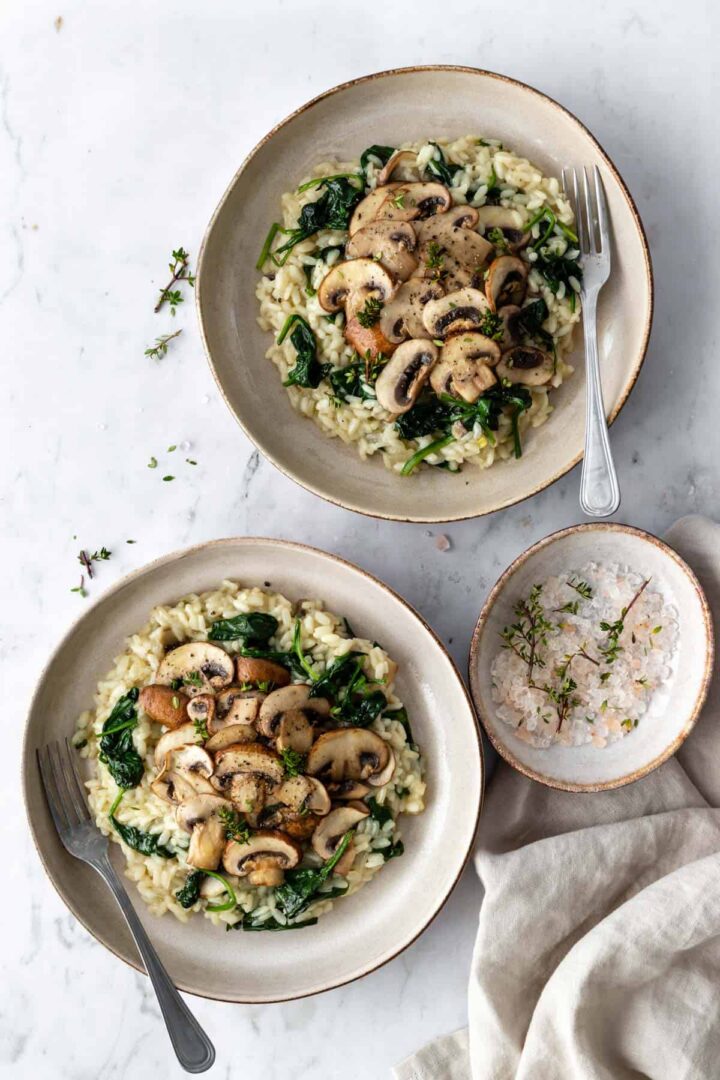 two plates with arborio rice, mushrooms, spinach, garnished with thyme and a fork on the side. A small bowl with salt and thyme on the side and a beige kitchen towel